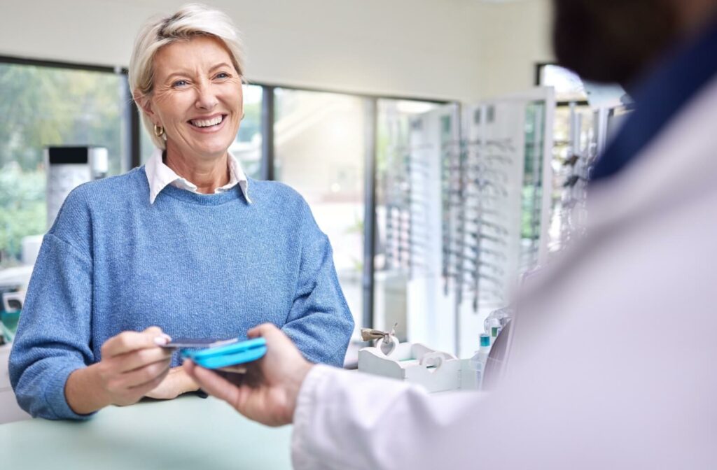 A smiling patient in a blue sweater pays for dry eye treatment in a well-lit and welcoming optometrist's office.