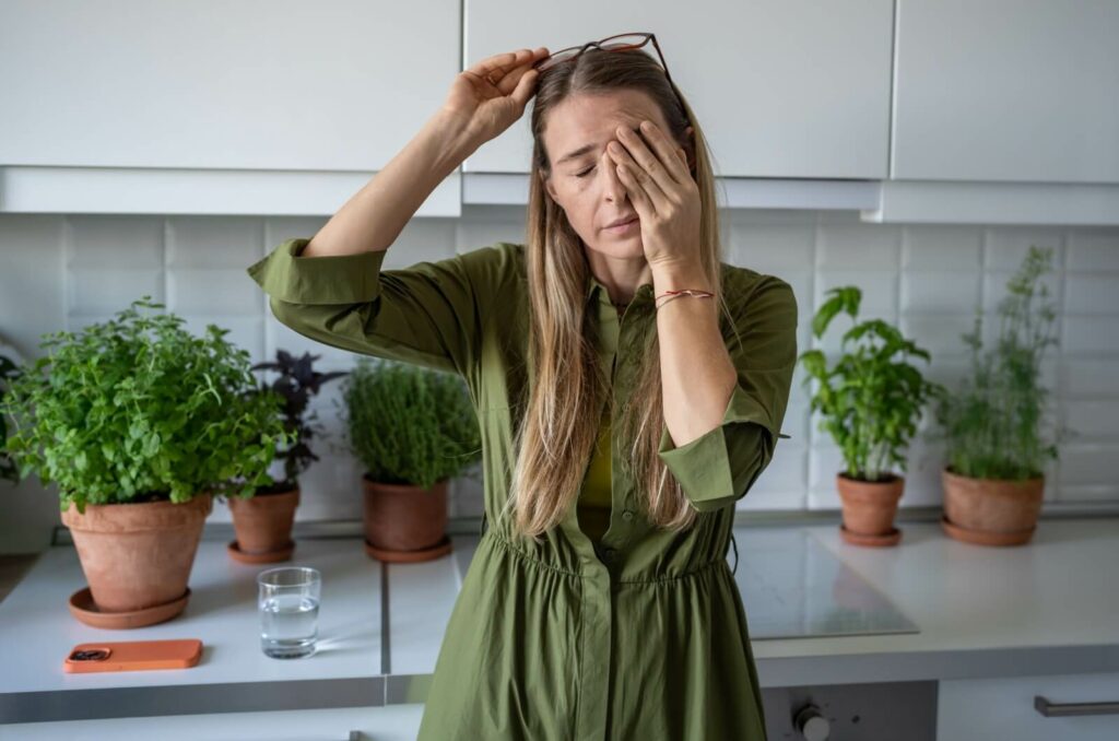 A person standing in their kitchen, rubbing their dry eyes.