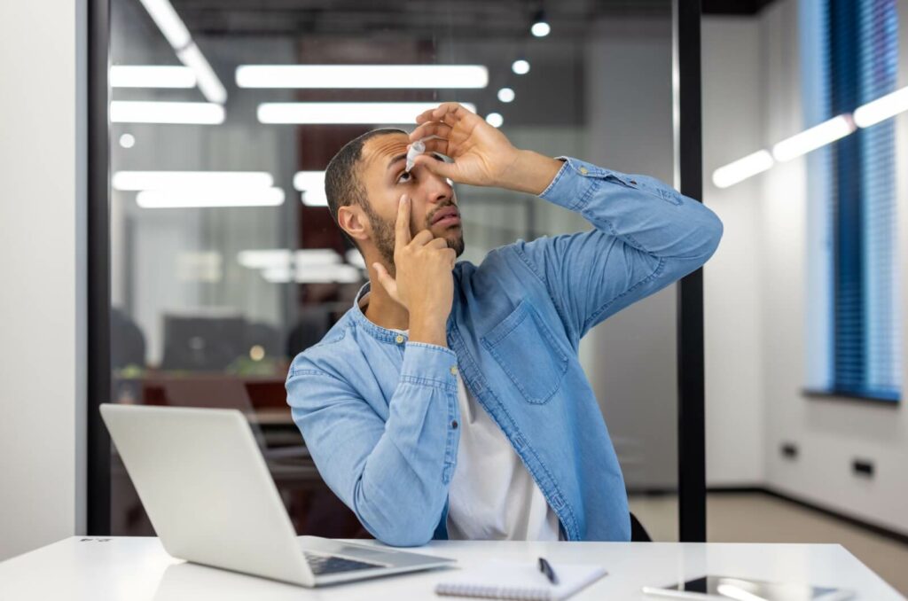 A business professional applies eye drops to their eyes while at work in an office cubicle