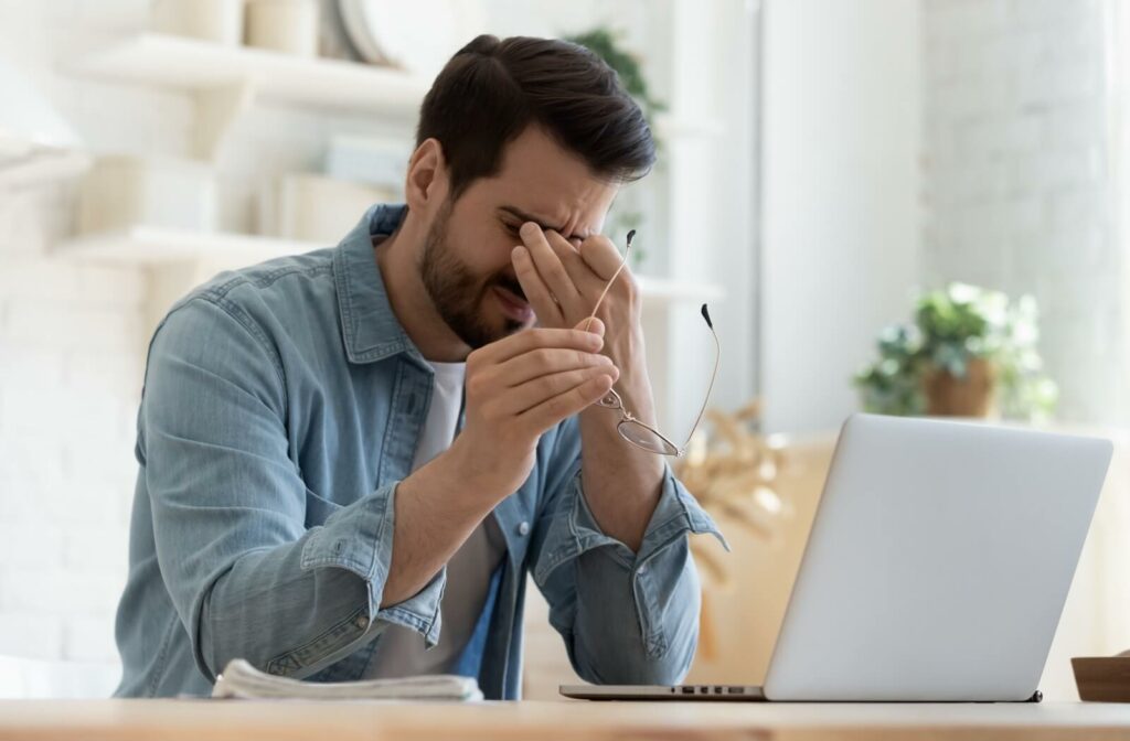 A person working at a laptop removes their glasses and rubs their dry eyes due to MGD.