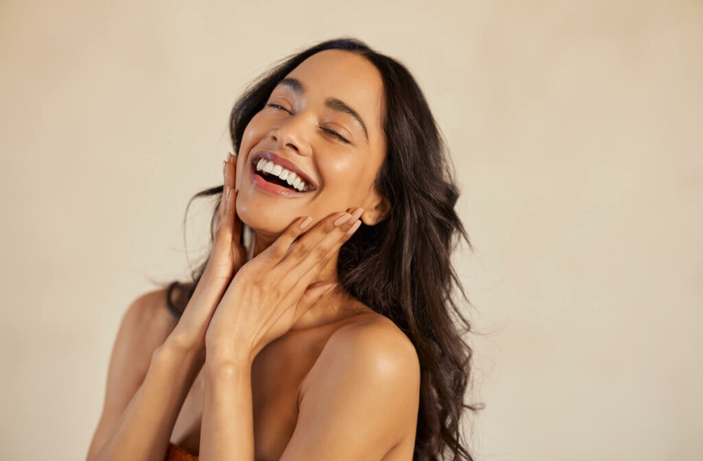 A woman with glowing skin smiles brightly while touching her face with both hands.