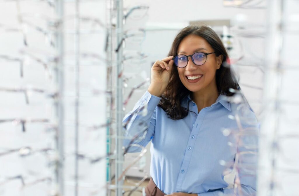 Smiling person wearing glasses and adjusting the frames while standing among rows of eyeglasses in an optical store.