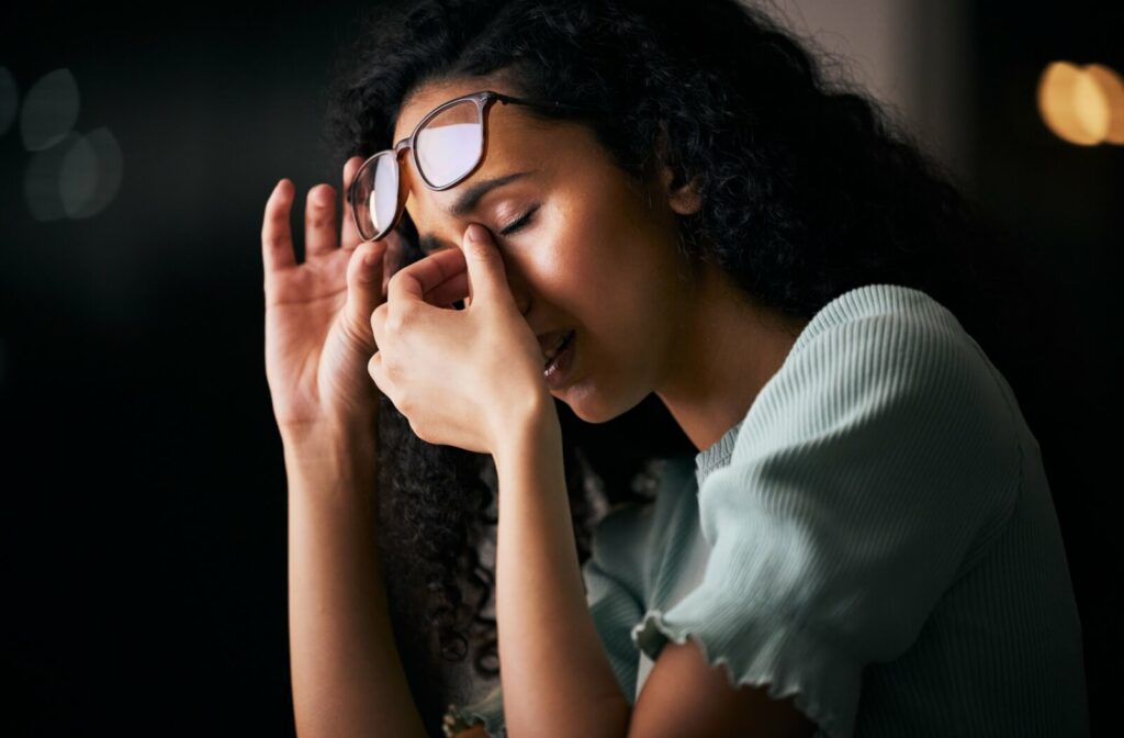 Individual experiencing eye strain or dry eye symptoms while removing glasses in low-light conditions.